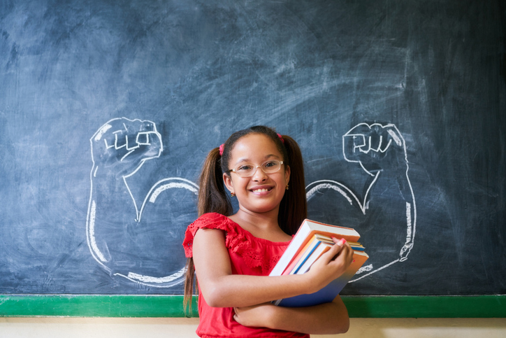 hispanic Girl Holding Books In Classroom And Smiling